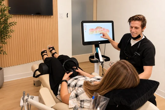 Smiling dentist showing a patient a digital 3D model of teeth on a monitor while she reclines in a dental chair.